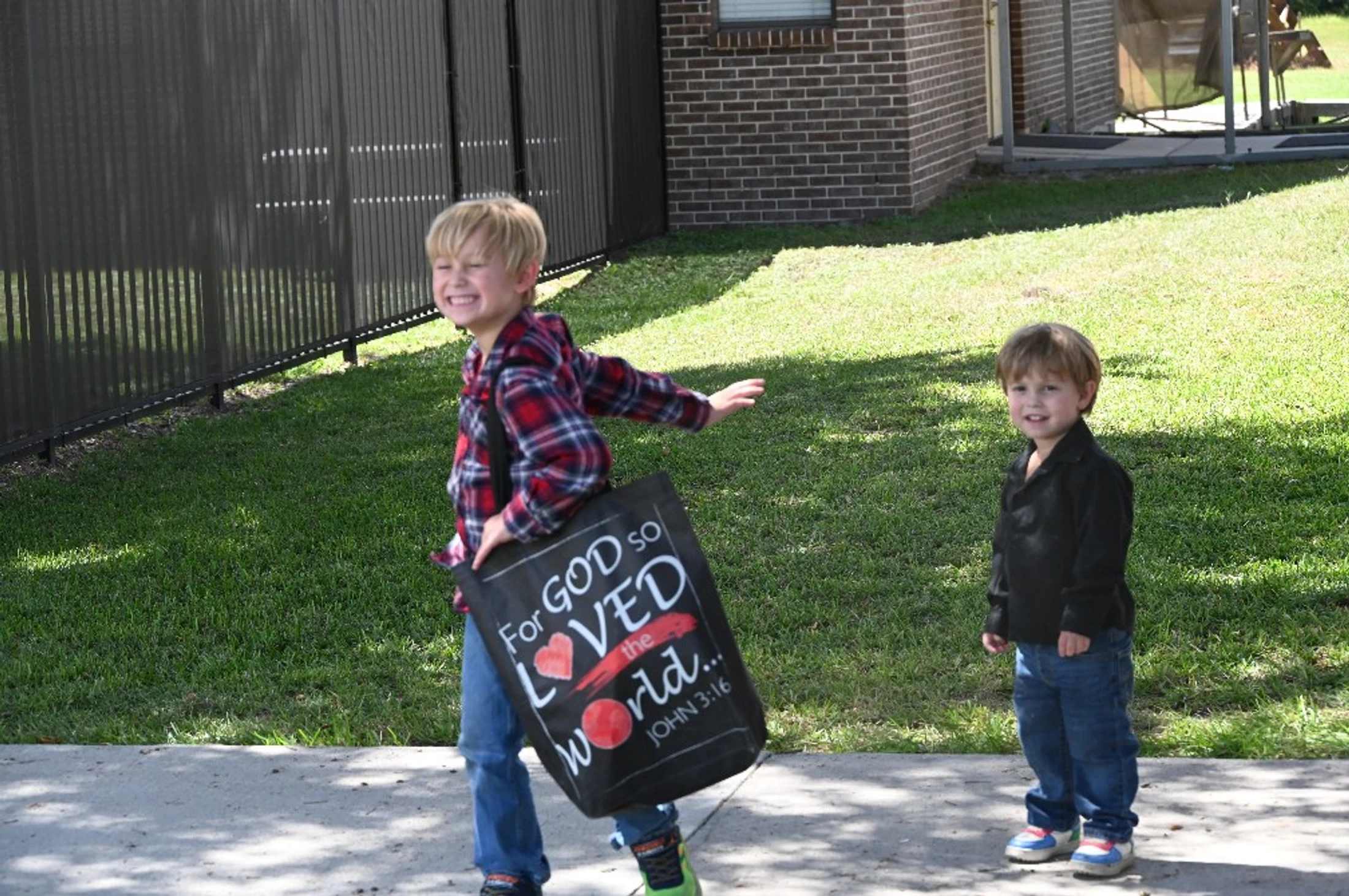 An older boy in a plaid shirt smiles as he walks carrying a black tote bag, while a younger boy in a black shirt stands on the grass nearby.